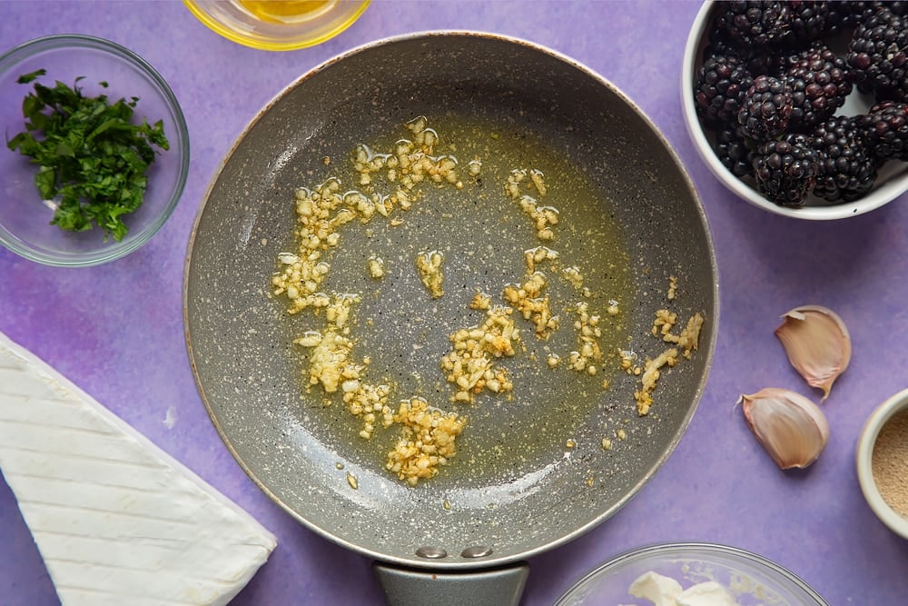 Fried garlic in a small frying pan. Ingredients for brie and blackberry pizza surround the pan.