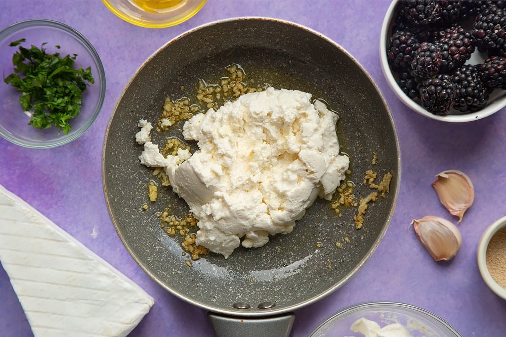 Fried garlic and ricotta in a small frying pan. Ingredients for brie and blackberry pizza surround the pan.