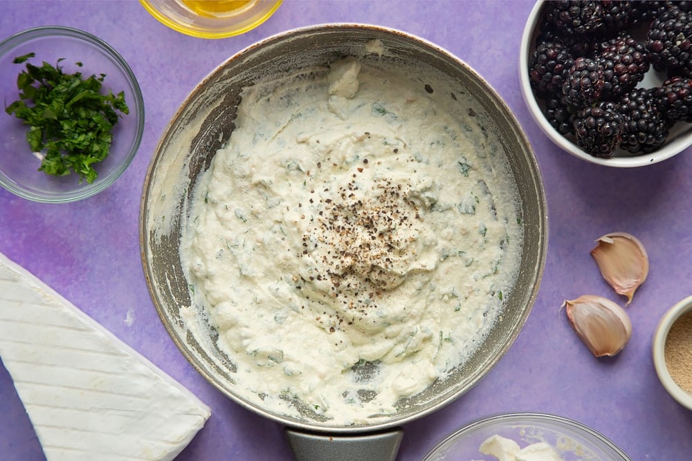 Ricotta whipped with garlic and parsley in a small frying pan, seasoned with pepper. Ingredients for brie and blackberry pizza surround the pan.