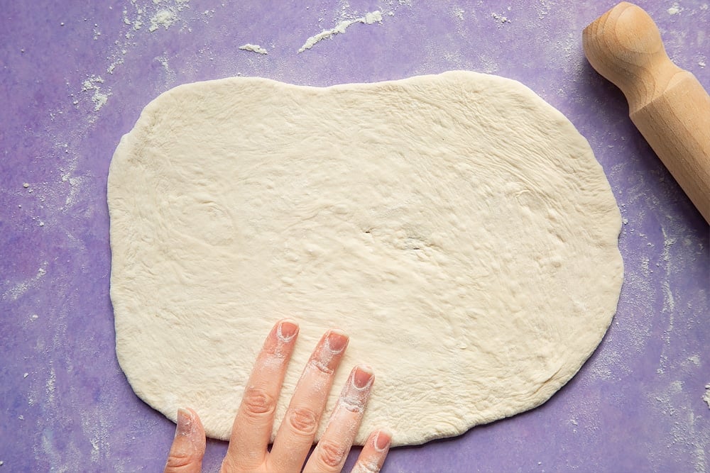 A rolled out oblong of pizza dough. Ingredients for brie and blackberry pizza surround the dough.