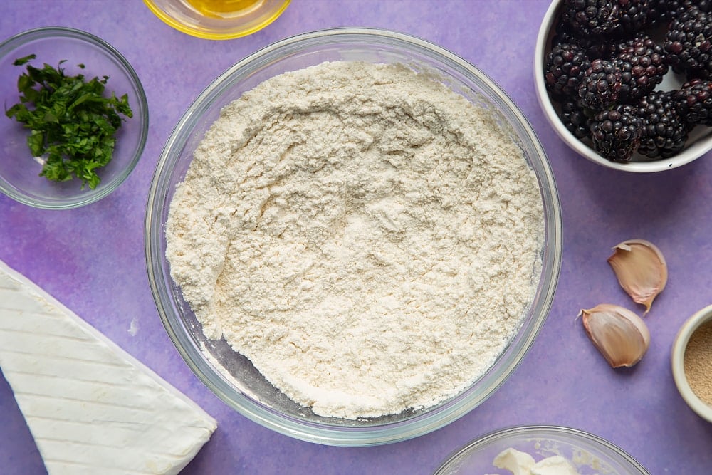 Flour, yeast and salt mixed together in a glass mixing bowl. Ingredients for brie and blackberry pizza surround the bowl.