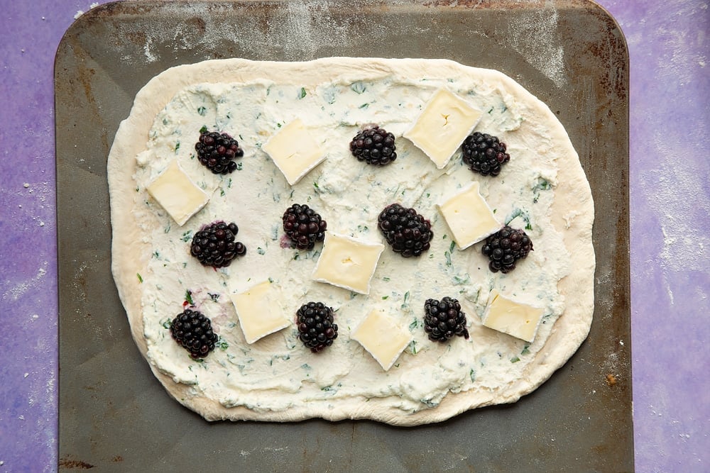 An oblong of pizza base on a baking sheet, spread with the ricotta mix and topped with blackberries and slices of brie.