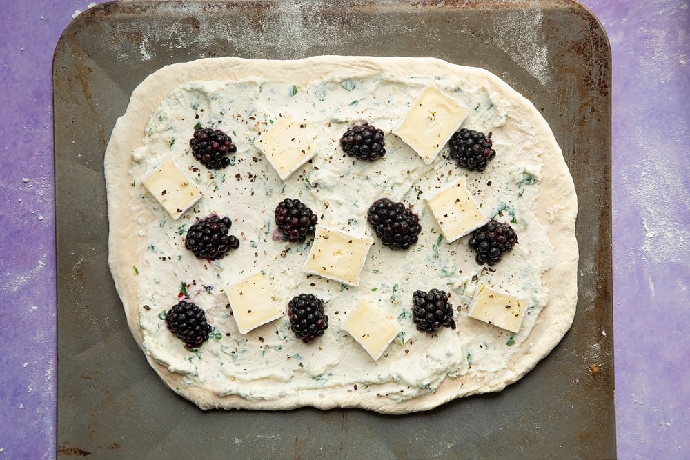 An oblong of pizza base on a baking sheet, spread with the ricotta mix and topped with blackberries and slices of brie. Seasoned with black pepper.