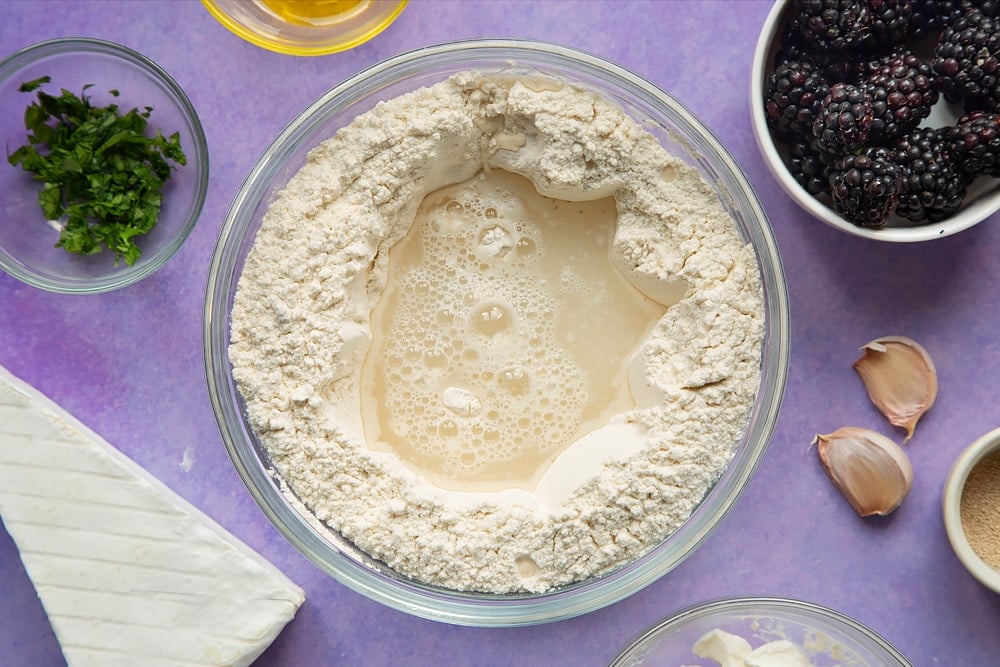 Flour, yeast and salt mixed together in a glass mixing bowl. Water is poured into a well in the centre. Ingredients for brie and blackberry pizza surround the bowl.