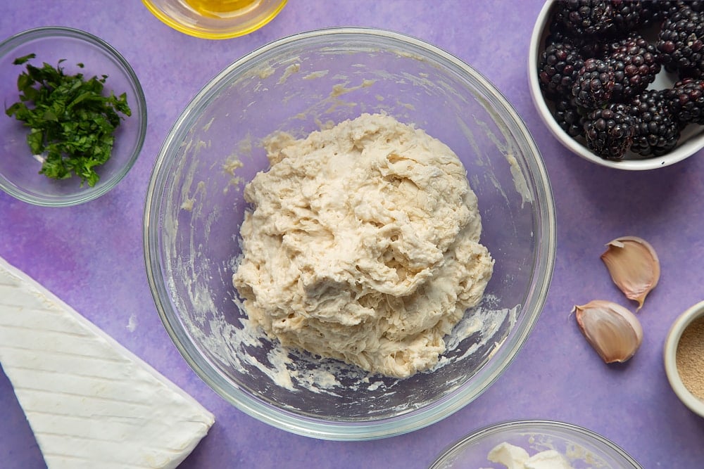 A rough pizza dough in a glass mixing bowl. Ingredients for brie and blackberry pizza surround the bowl.