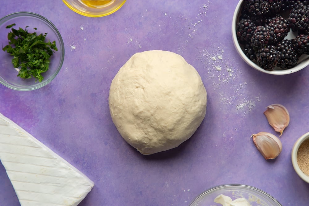 A smooth ball of kneaded pizza dough. Ingredients for brie and blackberry pizza surround the dough.