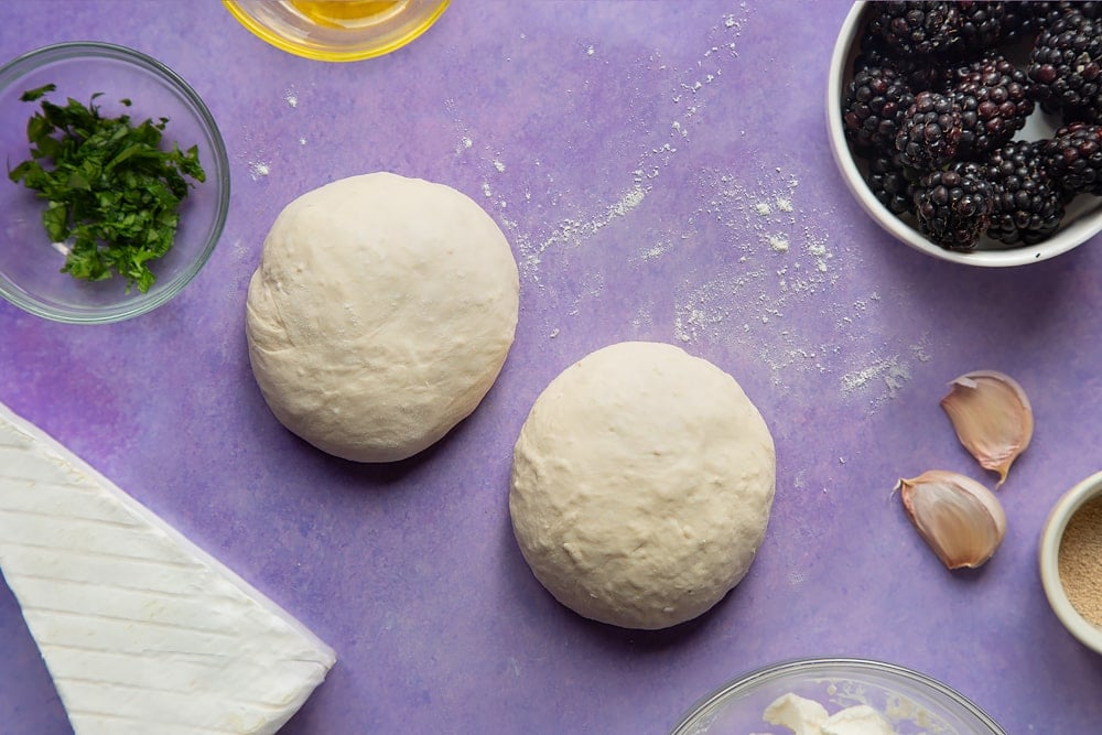 Two balls of pizza dough. Ingredients for brie and blackberry pizza surround the dough.