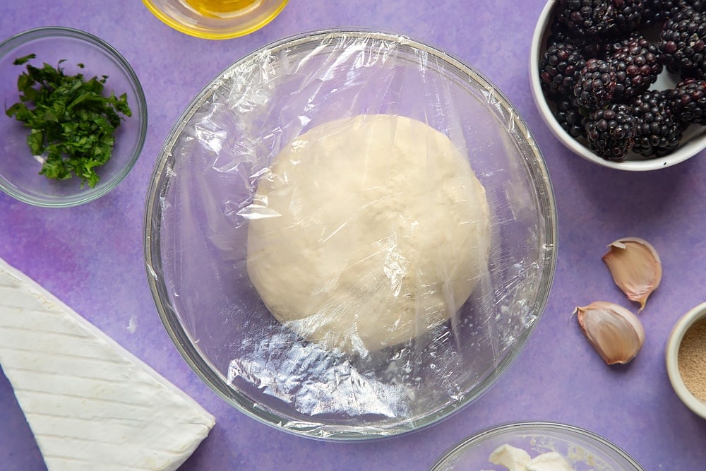 A smooth ball of kneaded pizza dough in a glass mixing bowl, covered with clingfilm. Ingredients for brie and blackberry pizza surround the bowl.