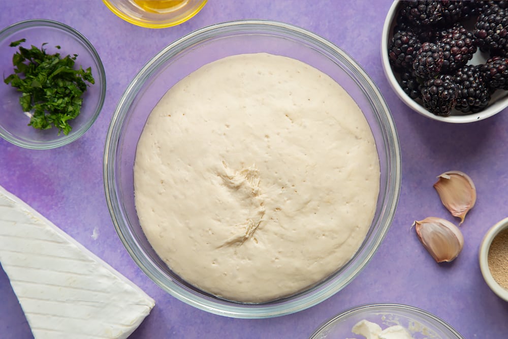 Proved pizza dough in a glass mixing bowl. Ingredients for brie and blackberry pizza surround the bowl.