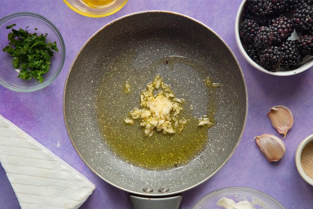 Oil and garlic in a small frying pan. Ingredients for brie and blackberry pizza surround the pan.