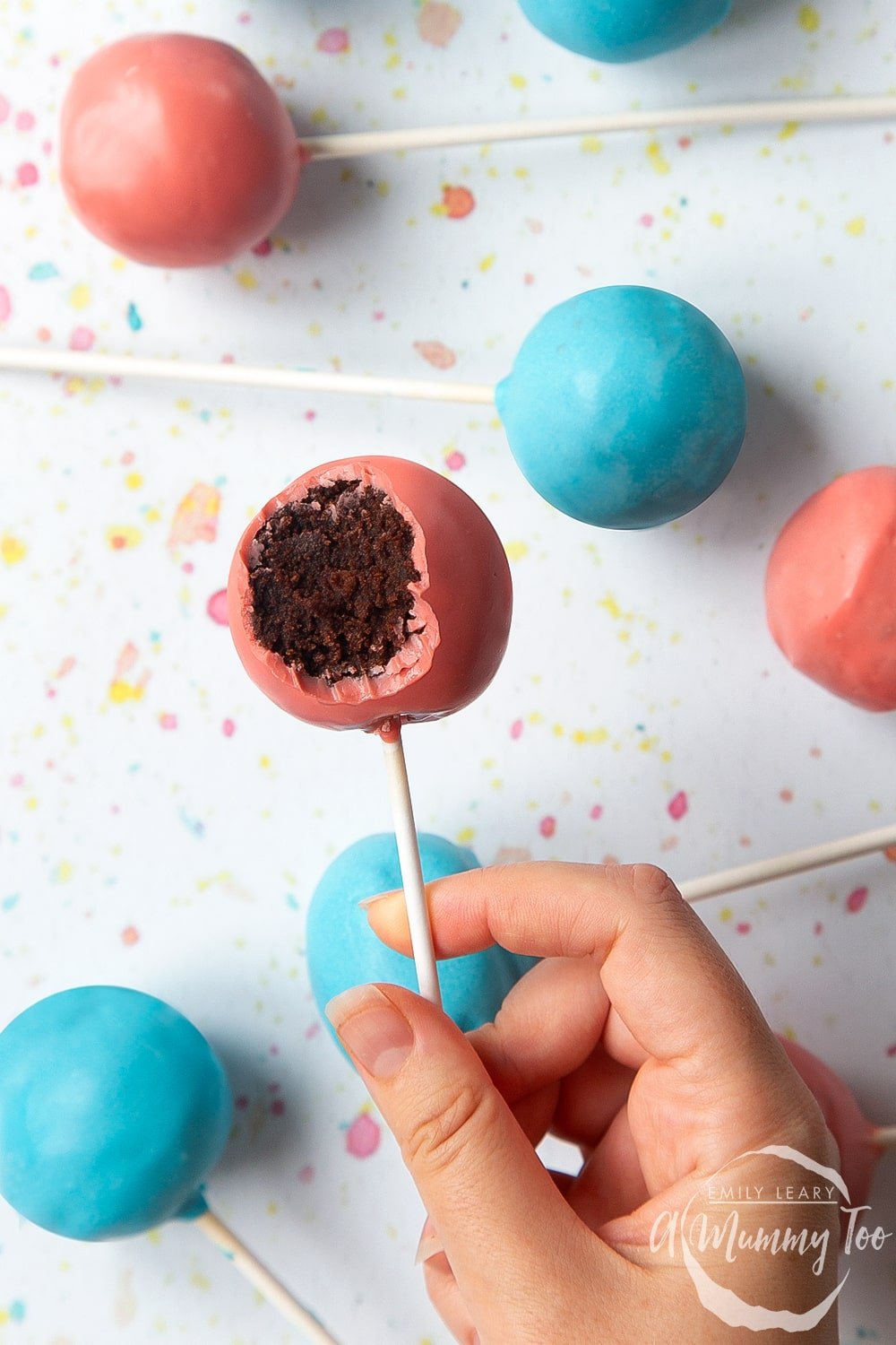 A hand holds a pink cake pop from a cake pop bouquet. It has a bite taken out of it, showing the chocolate sponge filling inside.