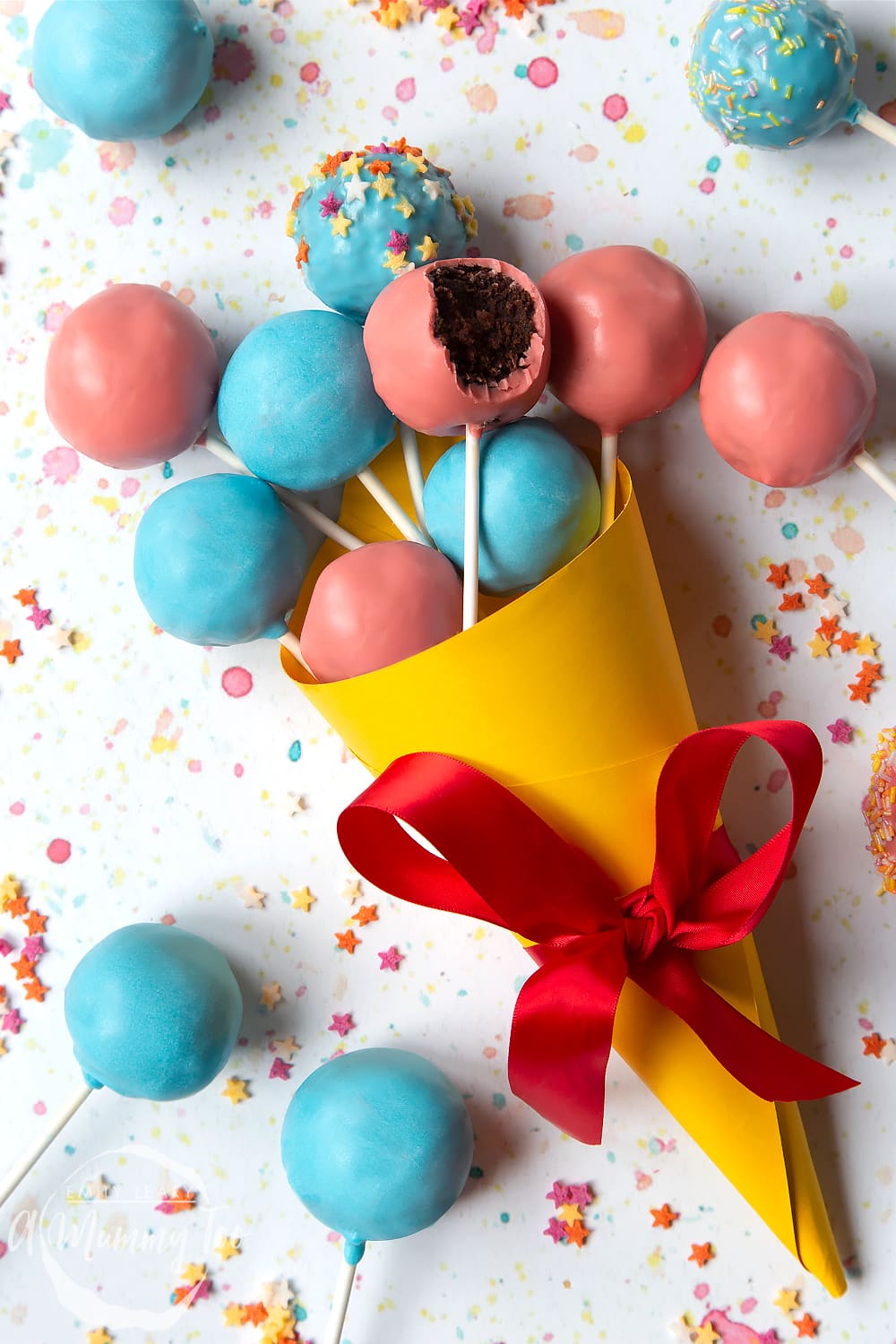 A cake pop bouquet surrounded by other cake pops. The bouquet is made from several blue and pink cake pops, gathered together and wrapped in a yellow paper cone with a red ribbon.