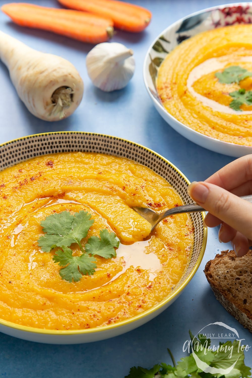 Bowl of carrot and parsnip soup in a decorative plate on a blue background. There's an additional bowl of the soup in the background alongside a garlic bulb and a parsnip.