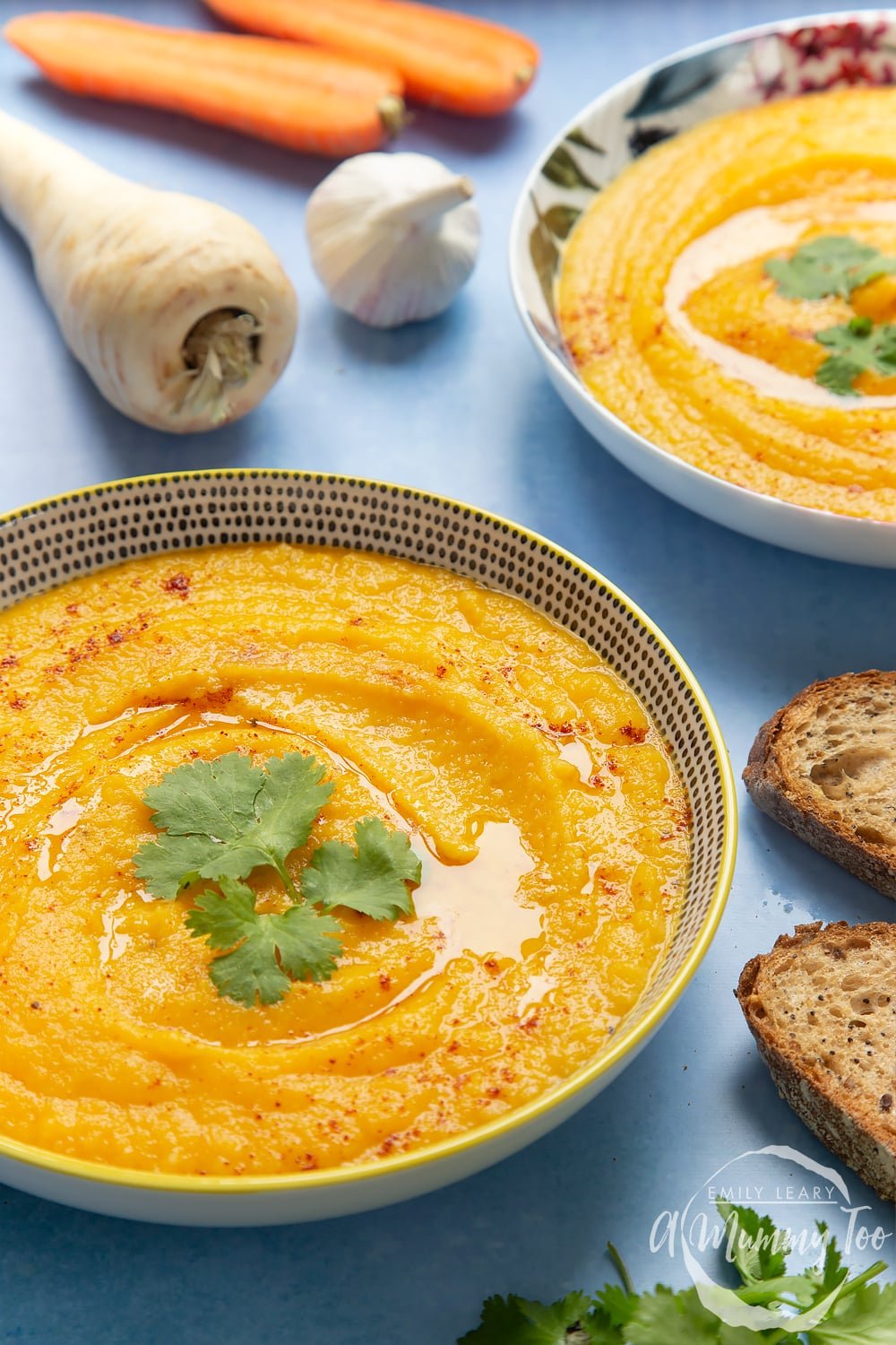 Two bowls of carrot and parsnip soup on a blue background. At the side there are two slices of bread, a parsnip some garlic and some carrots. 