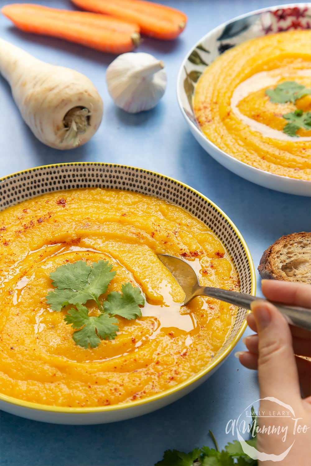 Two bowls of carrot and parsnip soup. A hand holding a spoon is going into one of the bowls. The sides are decorated with ingredients used in the carrot and parsnip soup recipe.