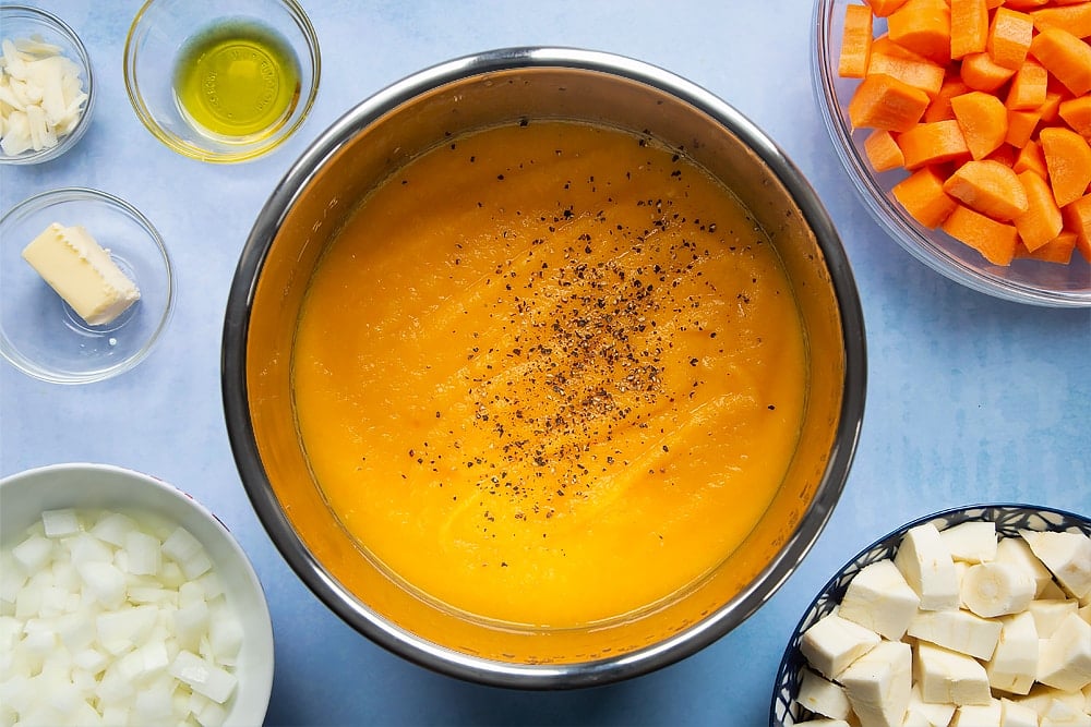 Pan of carrot and parsnip soup with added seasoning. At the side of there's a number of bowls filled with the soups ingredients.