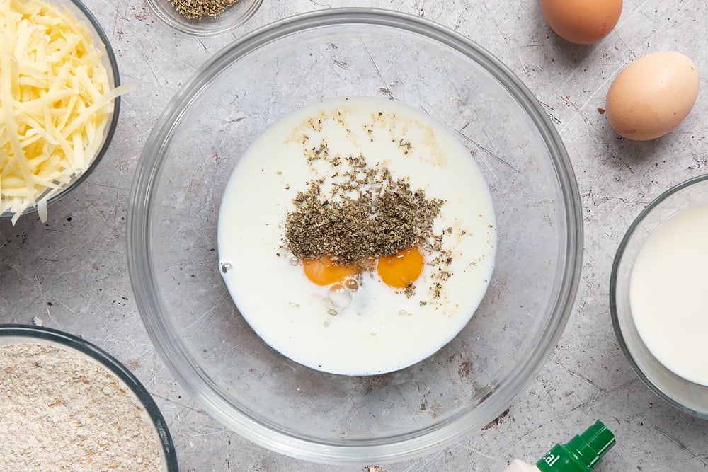 Eggs, milk, garlic granules and dried sage in a glass mixing bowl. Ingredients to make easy cheese muffins surround the bowl.