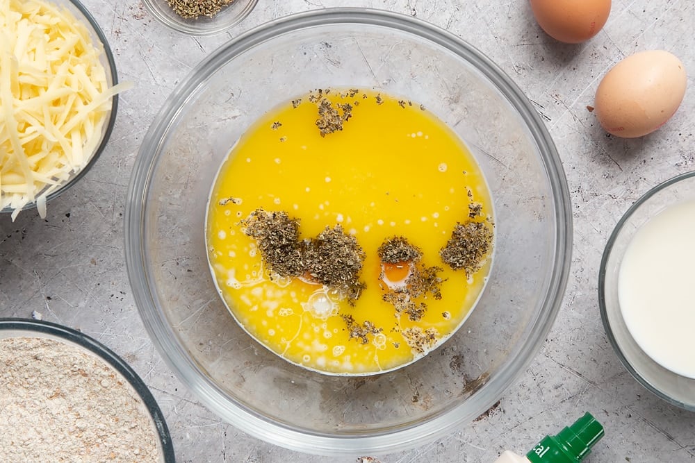 Eggs, milk, garlic granules, dried sage and melted butter in a glass mixing bowl. Ingredients to make easy cheese muffins surround the bowl.