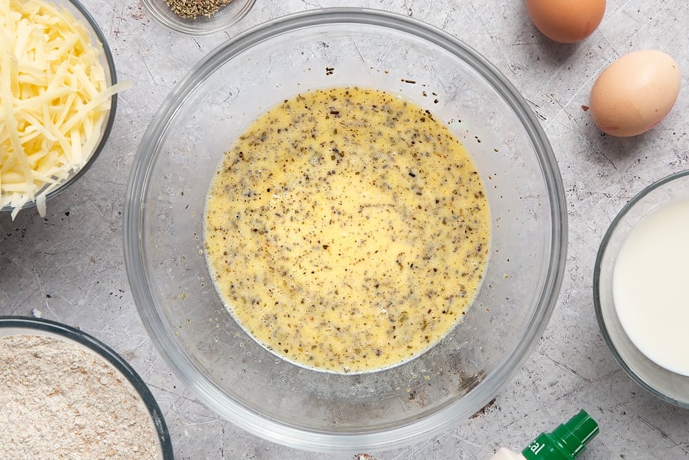 Eggs, milk, garlic granules, dried sage and melted butter whisked together in a glass mixing bowl. Ingredients to make easy cheese muffins surround the bowl.