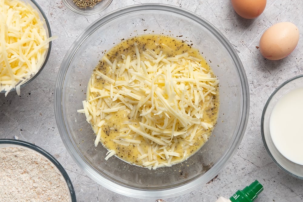 Eggs, milk, garlic granules, dried sage, melted butter and grated cheese in a glass mixing bowl. Ingredients to make easy cheese muffins surround the bowl.