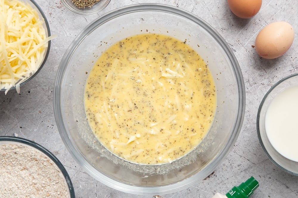 Eggs, milk, garlic granules, dried sage, melted butter and grated cheese mixed together in a glass mixing bowl. Ingredients to make easy cheese muffins surround the bowl.