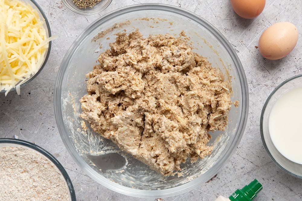 Easy cheese muffin dough in a glass mixing bowl. Ingredients to make easy cheese muffins surround the bowl.