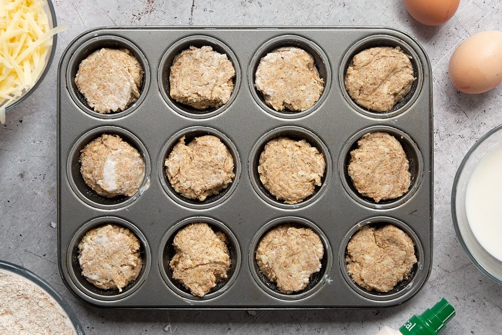 Easy cheese muffin dough in a 12-hole muffin tray. Ingredients to make easy cheese muffins surround the tray.