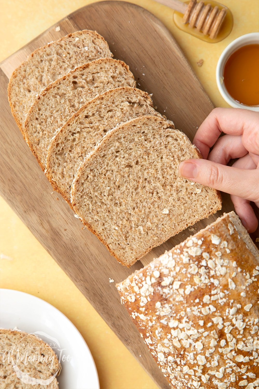 Front angle shot of a hand holding light honey and oat bread served on a wooden plate with a mummy too logo in the lower-left corner