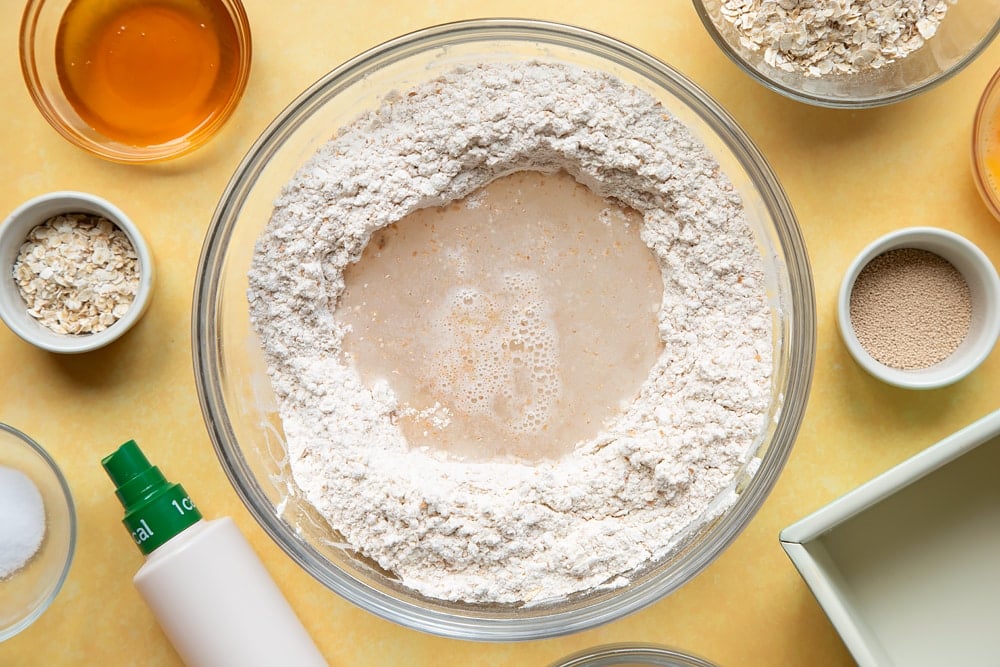 Overhead shot of mixed flour, oats, yeast, and salt with a well in the center in a large clear bowl