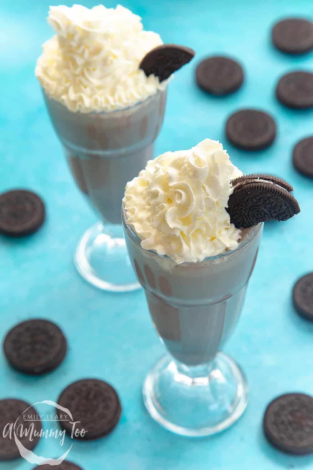 Two oreo milkshakes topped with squirty cream and half an oreo on a blue background. On the blue background there's also additional oreo cookies.