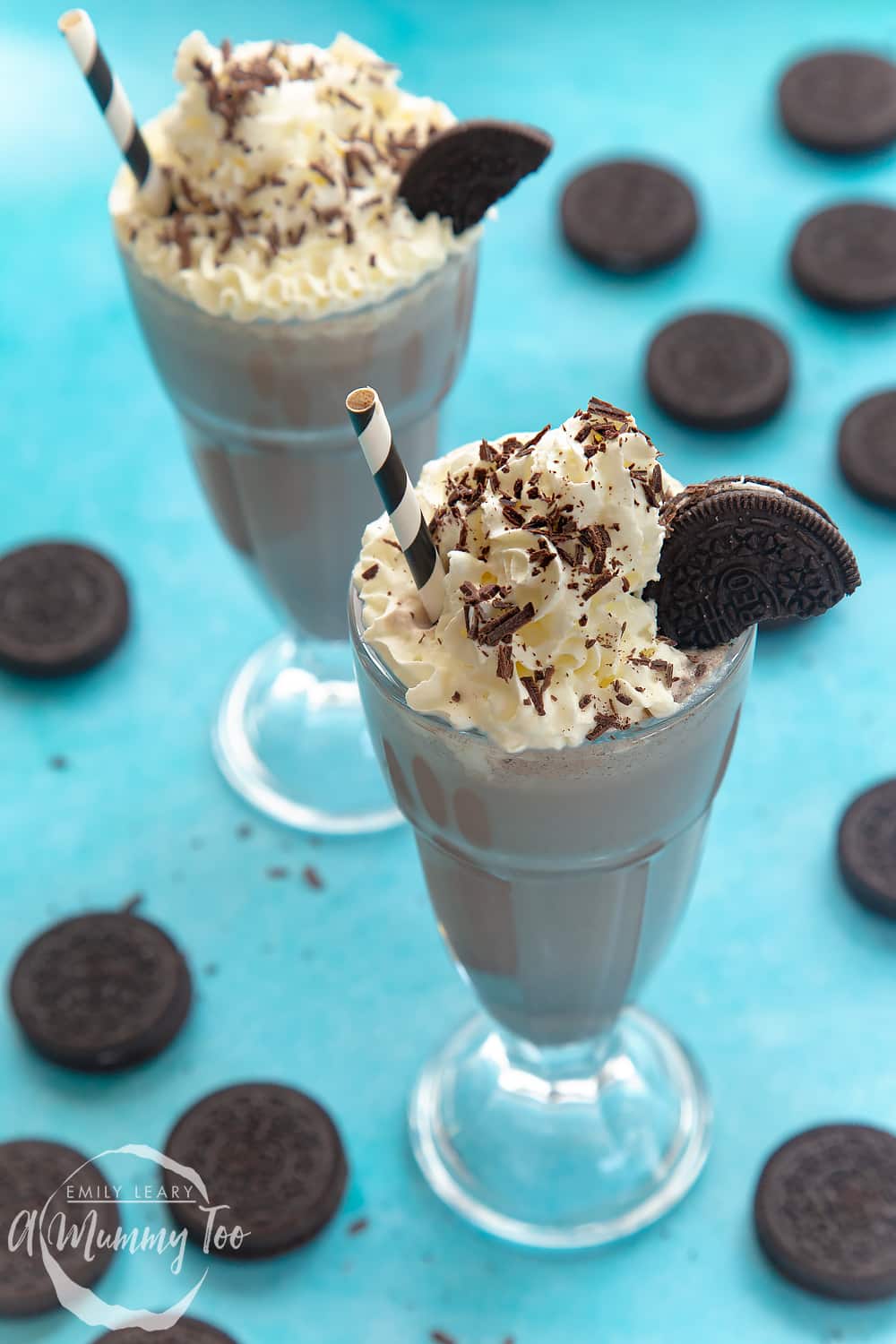 Two oreo milkshakes in a tall glass on a blue background. There's some additional oreos scattered around the milkshakes.