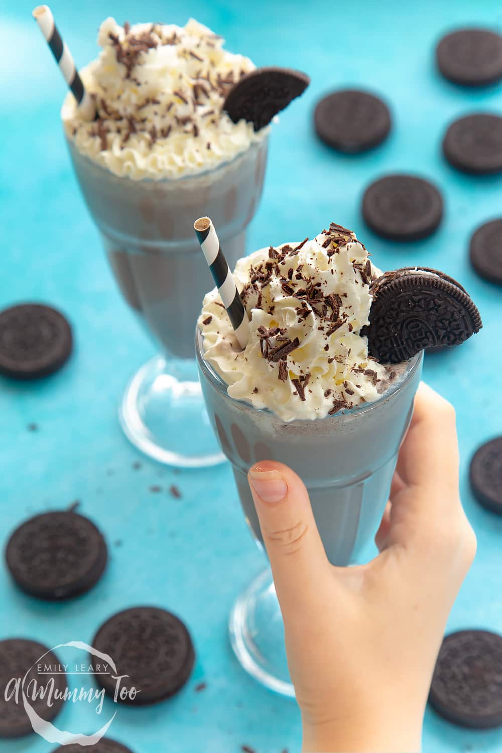 Two large milkshake glasses filled with oreo milkshake. In the background there are some additional oreos scattered around. 