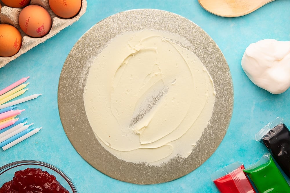 Vanilla buttercream spread thinly on a round silver cake board. Ingredients to make pizza cake surround the board.