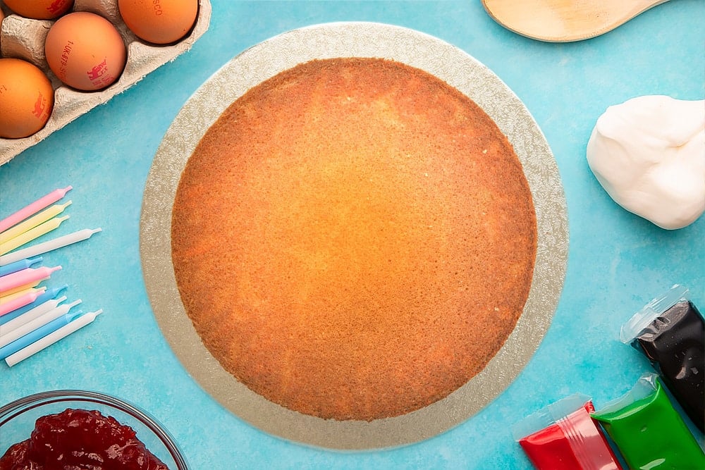 A vanilla sponge on a round silver cake board. Ingredients to make pizza cake surround the board.