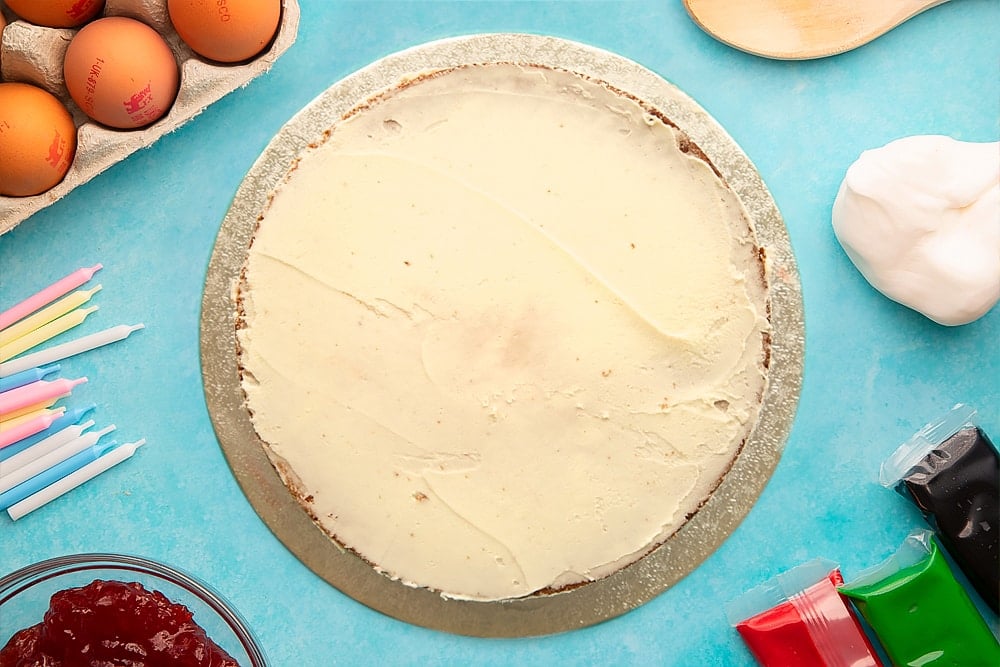 A vanilla sponge sandwich cake spread with buttercream on a silver cake board. Ingredients to make pizza cake surround the board.