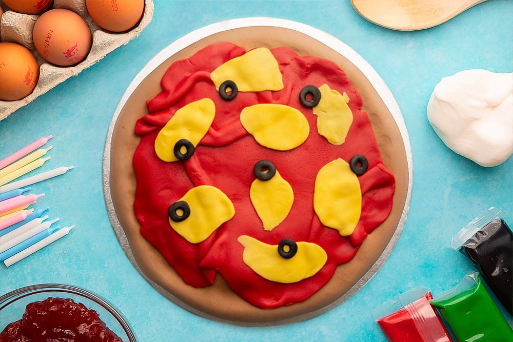 A vanilla sponge sandwich cake covered with brown, red, yellow and black sugar paste to resemble a pizza with olives on a silver cake board. Ingredients to make pizza cake surround the board.