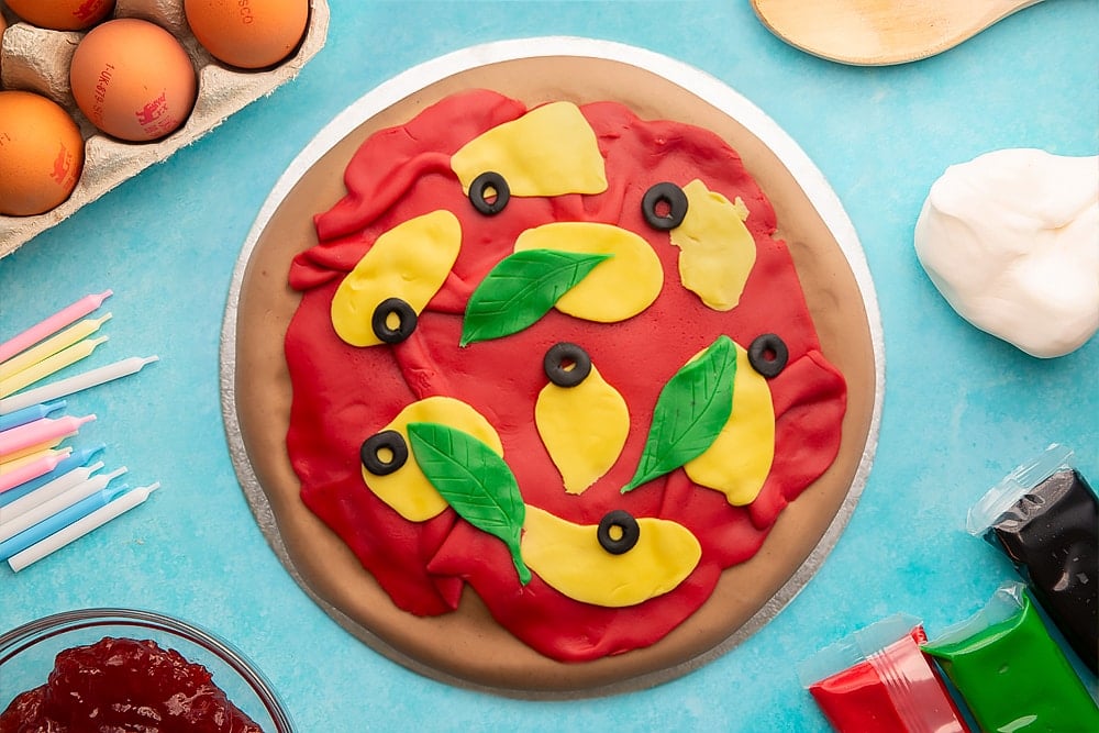 A vanilla sponge sandwich cake covered with brown, red, yellow, black and green sugar paste to resemble a pizza with olives and basil on a silver cake board. Ingredients to make pizza cake surround the board.