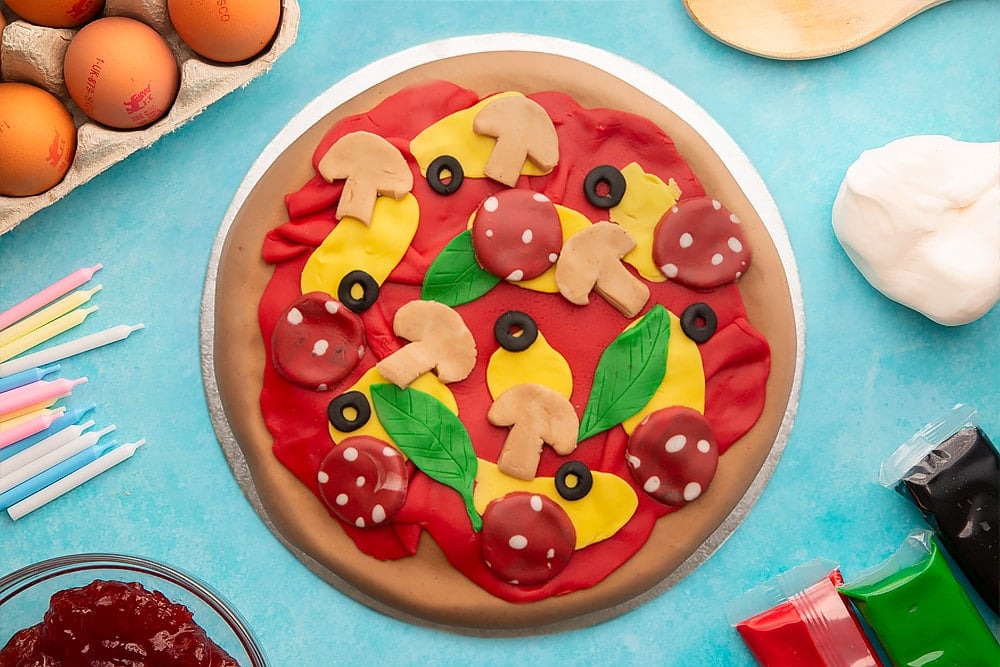 A vanilla sponge sandwich cake covered with brown, red, yellow, black, green and beige sugar paste to resemble a pizza with olives, basil. mushrooms and pepperoni on a silver cake board. Ingredients to make pizza cake surround the board.