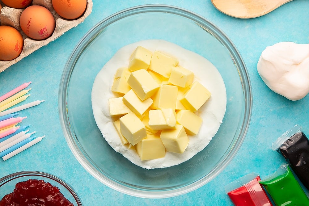 Sugar and cubed butter in a glass mixing bowl. Ingredients to make pizza cake surround the bowl.