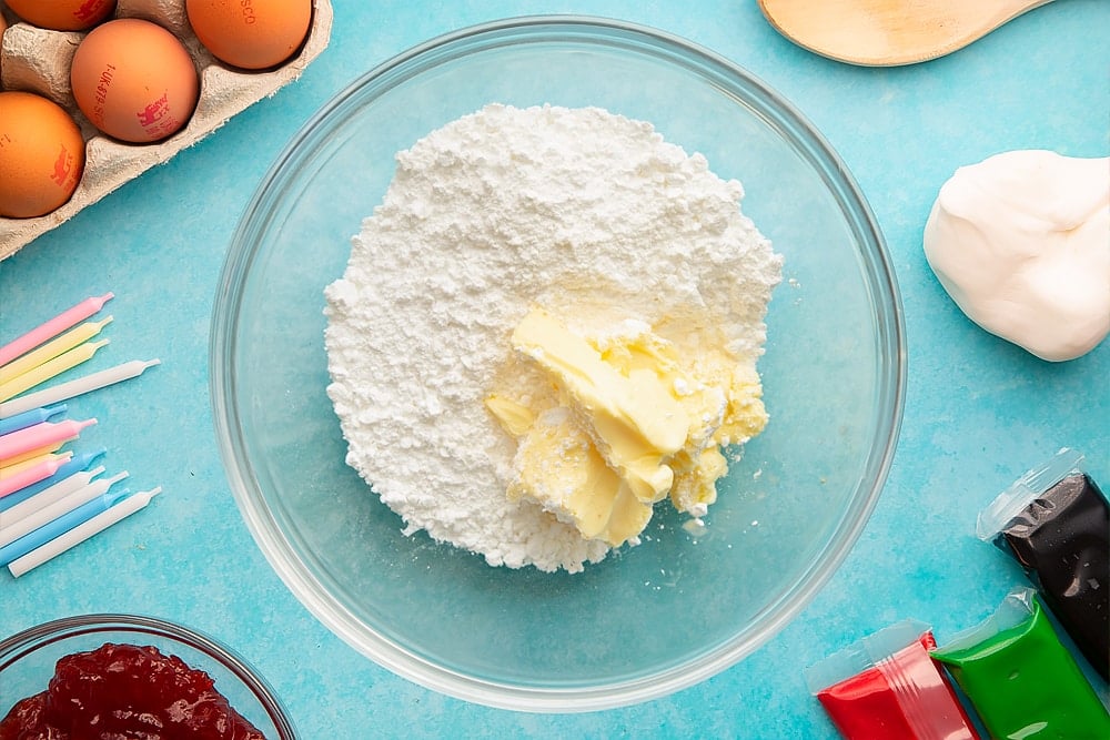 Vanilla, icing sugar and butter in a glass mixing bowl. Ingredients to make pizza cake surround the bowl.