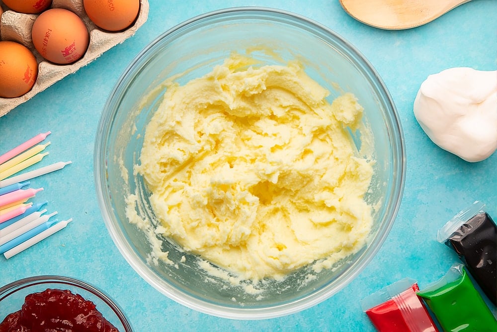 Sugar and butter creamed together in a glass mixing bowl. Ingredients to make pizza cake surround the bowl.