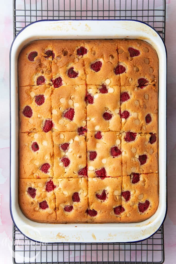A raspberry and white chocolate traybake in a white tray, baked and cooled on a wire rack and cut into 16 pieces. 