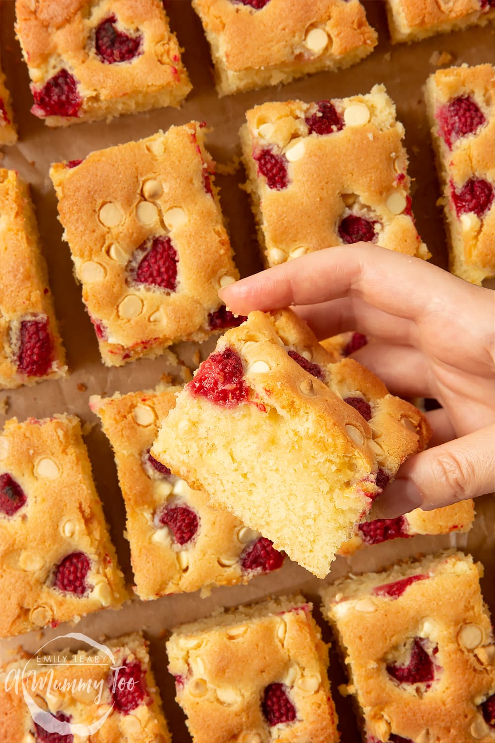 A slice of raspberry and white chocolate traybake held above a board with lots more slices on it.