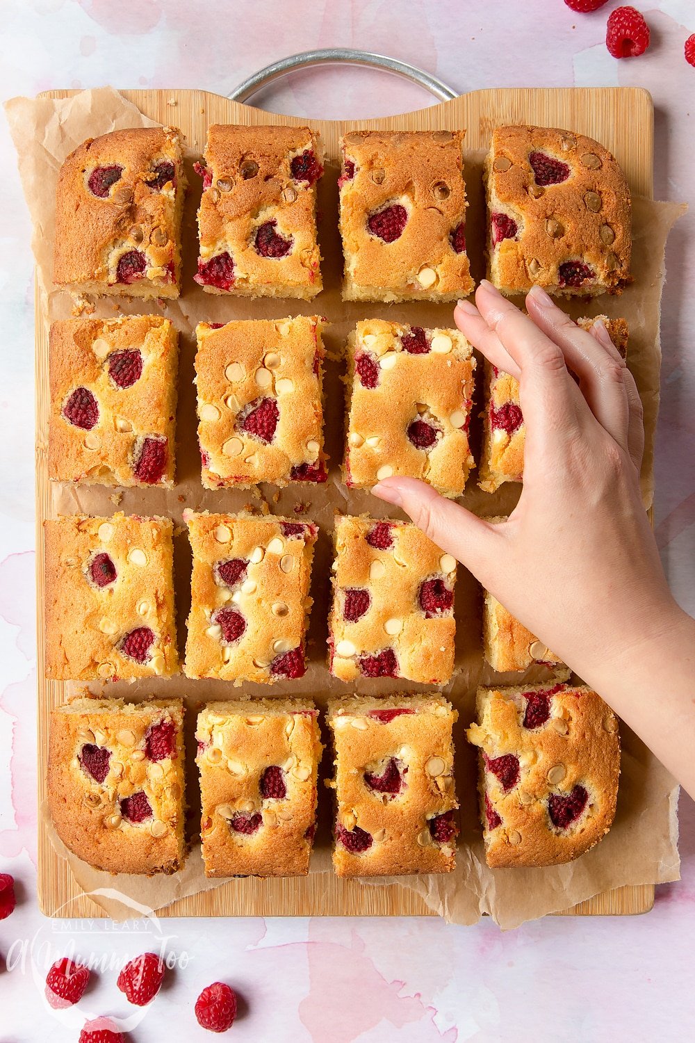 A wooden board with 16 pieces of raspberry and white chocolate traybake A hand reaches for a slice. 