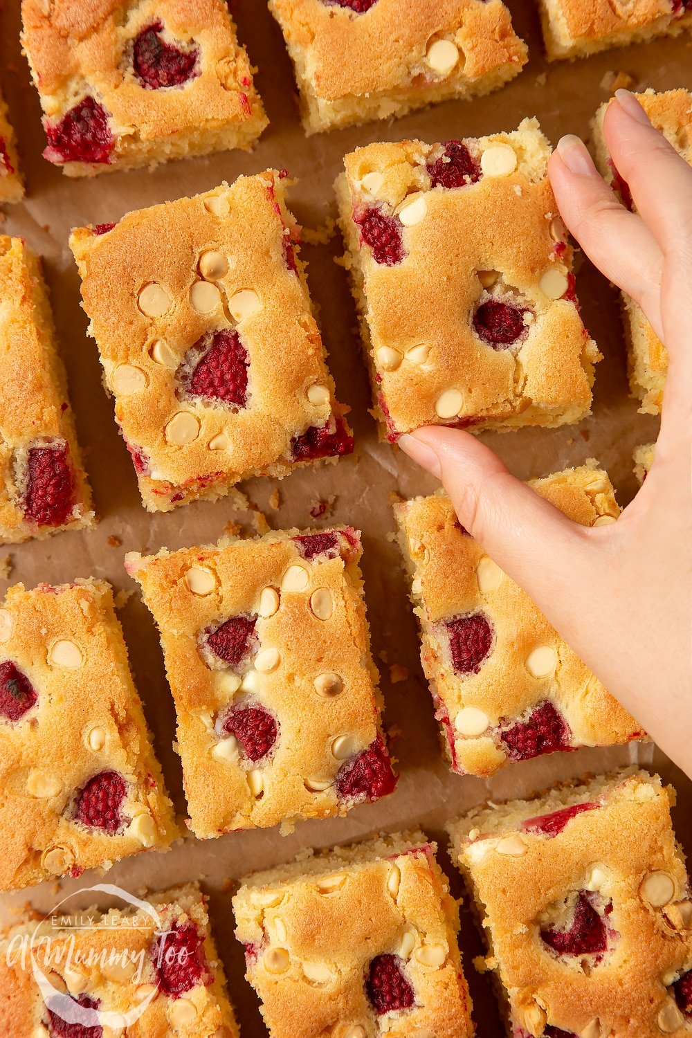 A hand picking up a piece of raspberry and white chocolate traybake. Slices are arranged on a wooden board.