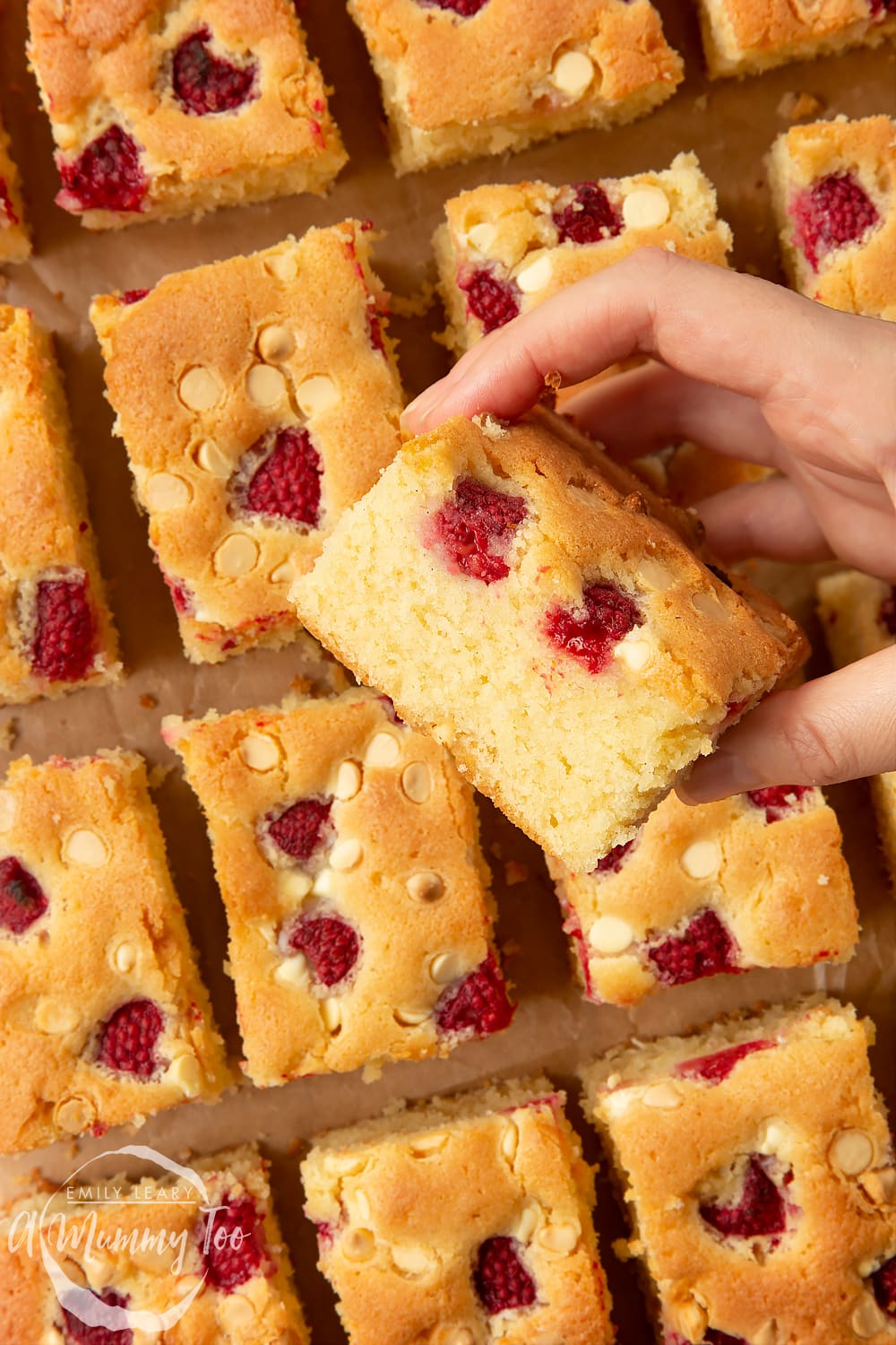 A raspberry and white chocolate traybake cut into 16 pieces. A hand holds a piece, showing the golden crumb inside.