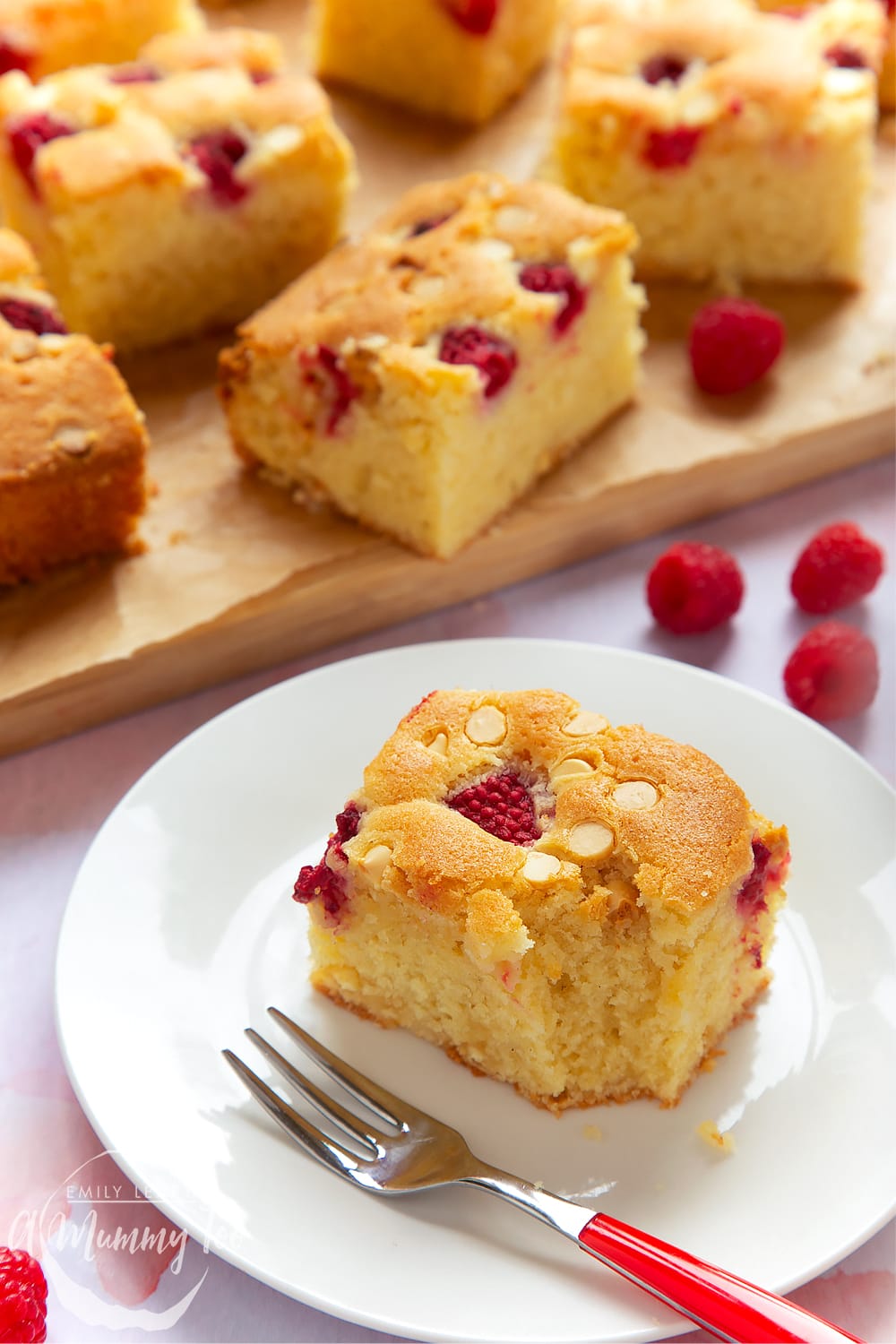 A piece of raspberry and white chocolate traybake served on a white plate. A red cake fork sits beside it.