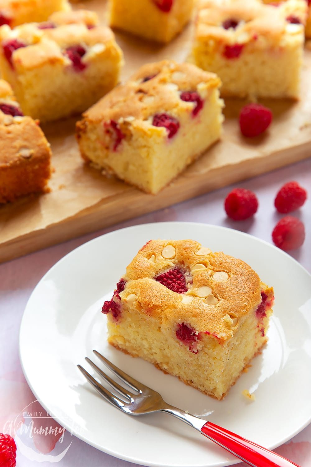 A piece of raspberry and white chocolate traybake served on a white plate. A red cake fork sits beside it. More slices are shown in the background.