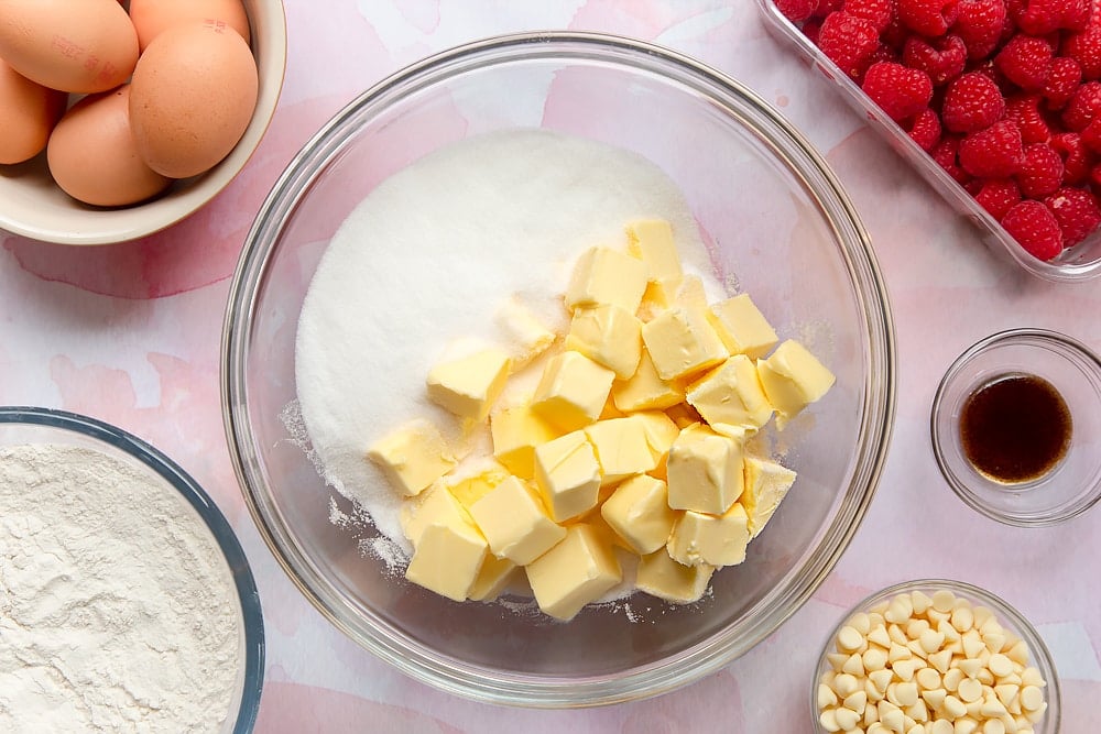 Sugar and butter in a bowl.Surrounding the bowl are ingredients to make a raspberry and white chocolate traybake