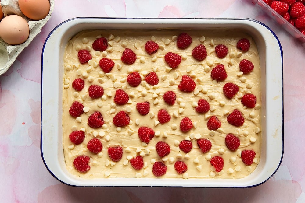 Cake batter with spread in a tray with white chocolate chips and fresh raspberries scattered on top. Surrounding the tray are ingredients to make a raspberry and white chocolate traybake